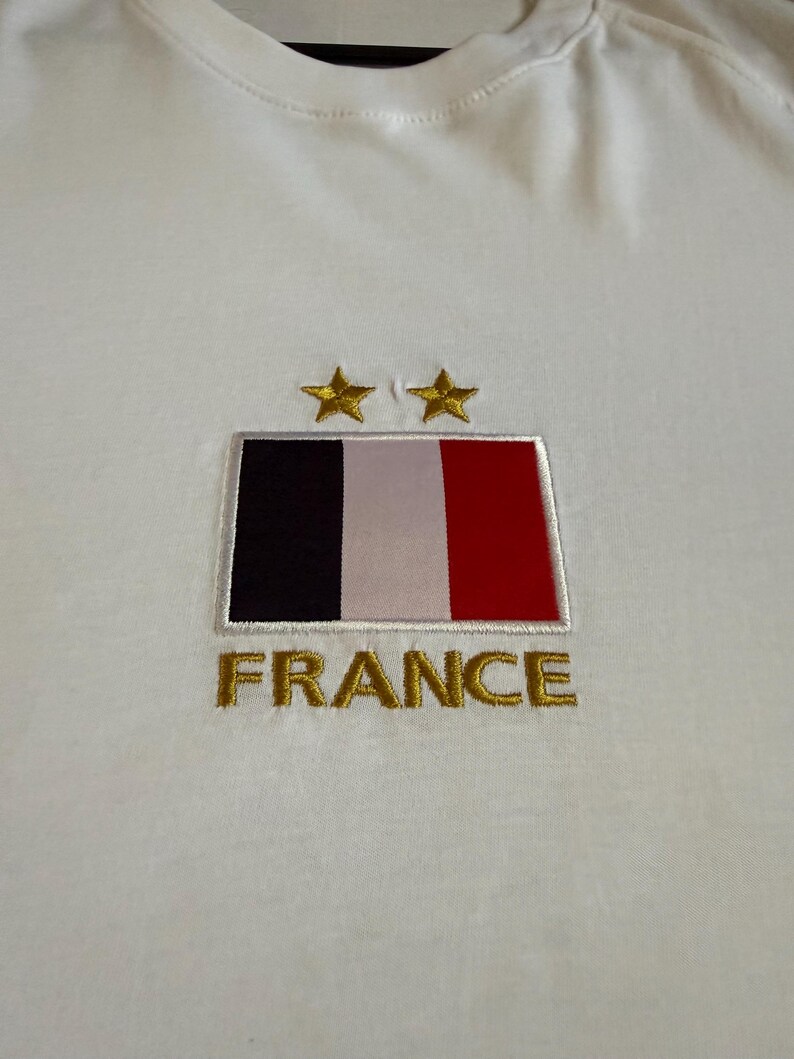 May include: A white t-shirt featuring an embroidered rectangular patch of the French flag with vertical black, white, and red stripes. Above the flag are two gold embroidered stars, and below the flag is the word FRANCE in gold embroidery.