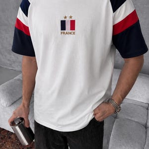 May include: A white short-sleeve t-shirt featuring raglan sleeves with navy blue and red stripes. The chest displays a rectangular French flag emblem with two gold stars above it and the word FRANCE embroidered in gold lettering below the flag.