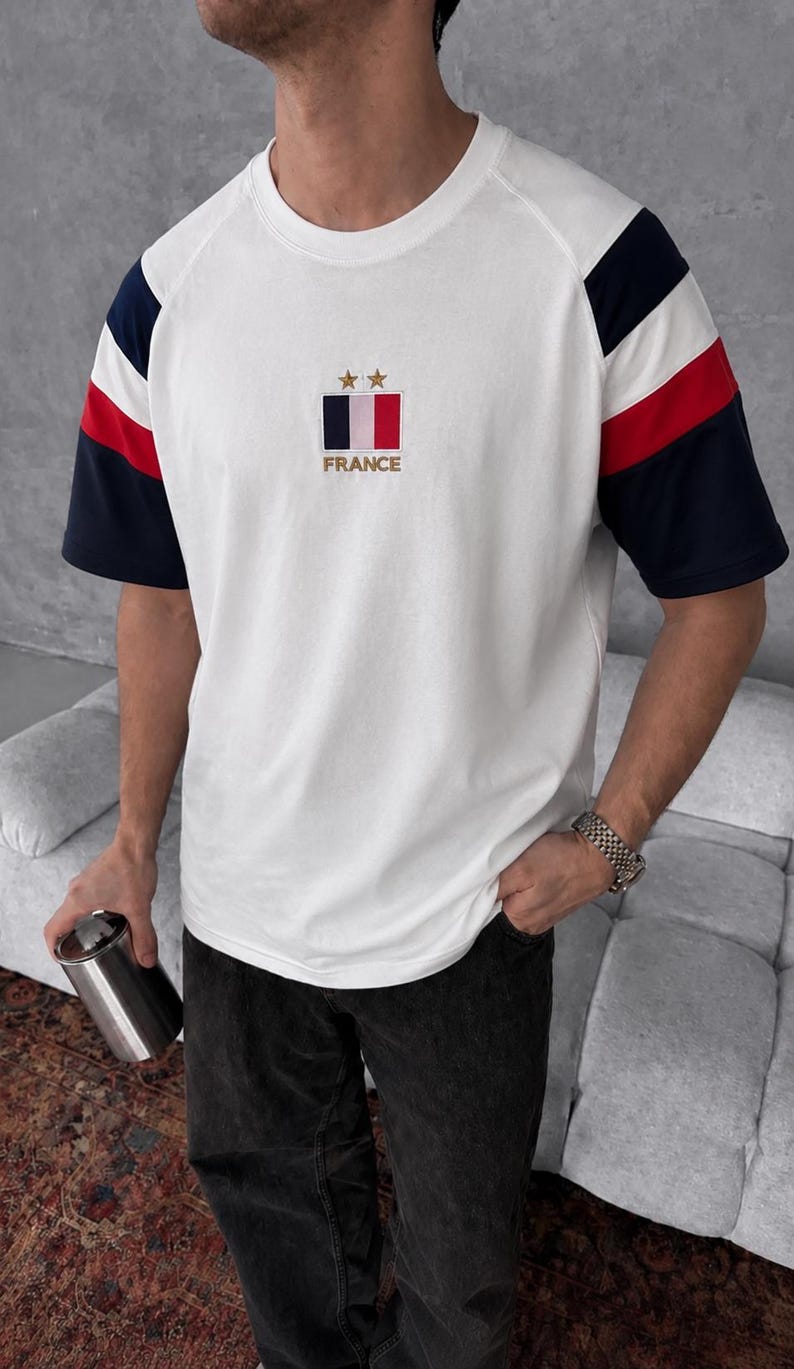 May include: A white short-sleeve t-shirt featuring raglan sleeves with navy blue and red stripes. The chest displays a rectangular French flag emblem with two gold stars above it and the word FRANCE embroidered in gold lettering below the flag.