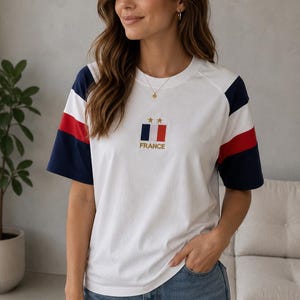 May include: A white short-sleeve t-shirt featuring navy blue and red stripes on the sleeves. The chest displays a small rectangular patch with the French flag, two gold stars above it, and the word FRANCE embroidered in gold lettering below.