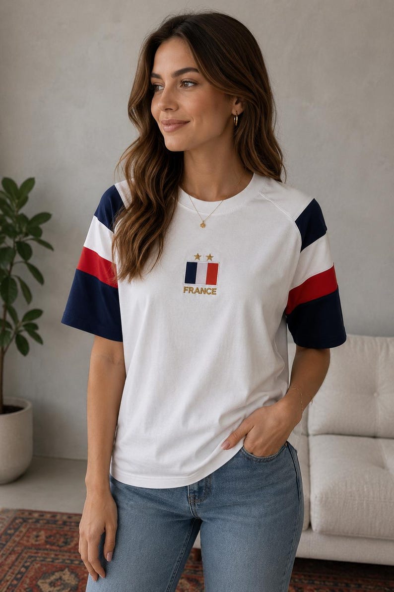 May include: A white short-sleeve t-shirt featuring navy blue and red stripes on the sleeves. The chest displays a small rectangular patch with the French flag, two gold stars above it, and the word FRANCE embroidered in gold lettering below.