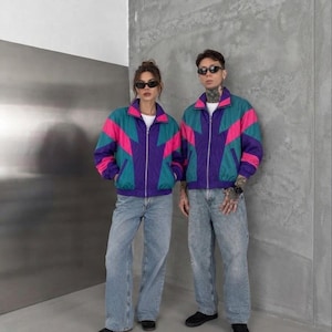 Peut inclure: Deux vestes de survêtement zippées assorties présentant un design color-block en bleu sarcelle, rose vif et violet foncé. Chaque veste possède un col montant, des manches longues et une fermeture éclair argentée, portées avec un jean bleu clair.