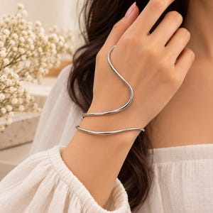 May include: A silver-toned metal hand bracelet featuring a continuous, fluid, wavy design that wraps around the wrist and extends upward across the back of the hand in a single, sleek, polished piece of jewelry with a smooth, reflective surface.