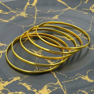 Könnte beinhalten: Fünf goldfarbene Armreifen liegen auf einem dunkelgrauen Teller mit goldfarbenen Adern. Die dünnen, kreisförmigen Armreifen reflektieren das Licht. Der Schmuck ist schlicht und elegant.