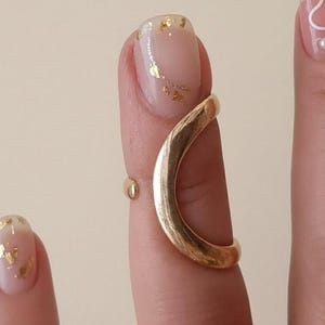 Puede incluir: Un anillo de dedo dorado con un diseño curvo, usado en un dedo con una uña rosa claro. La uña tiene detalles de hojas doradas. También se ve un pequeño anillo dorado en el mismo dedo. El fondo es de color beige neutro.