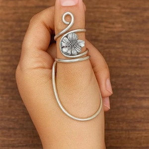 May include: A silver-toned ring with a floral design on the finger. The ring wraps around the finger with a decorative flower and a curved band. The ring is worn on a hand with a wooden background.