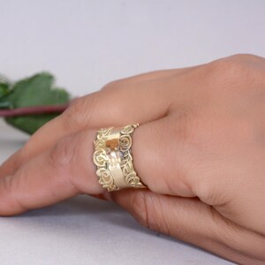 May include: A gold ring featuring an elaborate floral pattern. The ring is broad, with detailed scrollwork and a textured finish. The ring is worn on a finger, highlighting the intricate craftsmanship and the warm, metallic colour of the jewellery.