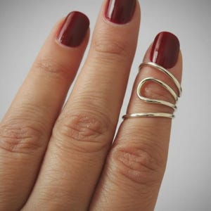 May include: A silver-colored finger ring with a unique, flowing design. The ring wraps around the finger with multiple bands and curves. The nails are painted a deep red color.