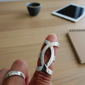 May include: A silver ring and a silver finger ring are displayed. The finger ring has a unique design with multiple bands that wrap around the finger. The other ring is a simple band. The background includes a wooden surface, a tablet, a cup, and a notepad.