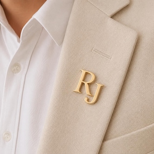 May include: A gold-toned metal brooch featuring the capital letters R and J in a serif font. The brooch is pinned to the lapel of a beige textured blazer worn over a crisp white collared shirt with visible buttons on the front placket.