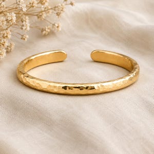 Könnte beinhalten: Ein goldfarbenes Armband in Manschettenform mit einer gehämmerten, strukturierten Oberfläche. Das Armband ist ein offener, kreisförmiger Reif mit abgerundeten, glatten Enden und einer reflektierenden Metalloberfläche, die das Licht auf der unebenen Struktur bricht.