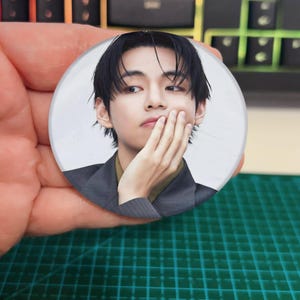 May include: A circular badge displaying a portrait of a person with dark hair, touching their face. The badge is held in a hand, with a green cutting mat and a keyboard in the background.