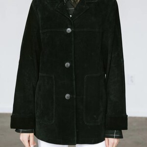 May include: A black suede jacket with a classic design. The jacket features a collared neckline, three buttons, and two front pockets. The sleeves have a cuff detail. The model is wearing a black and white patterned shirt underneath.