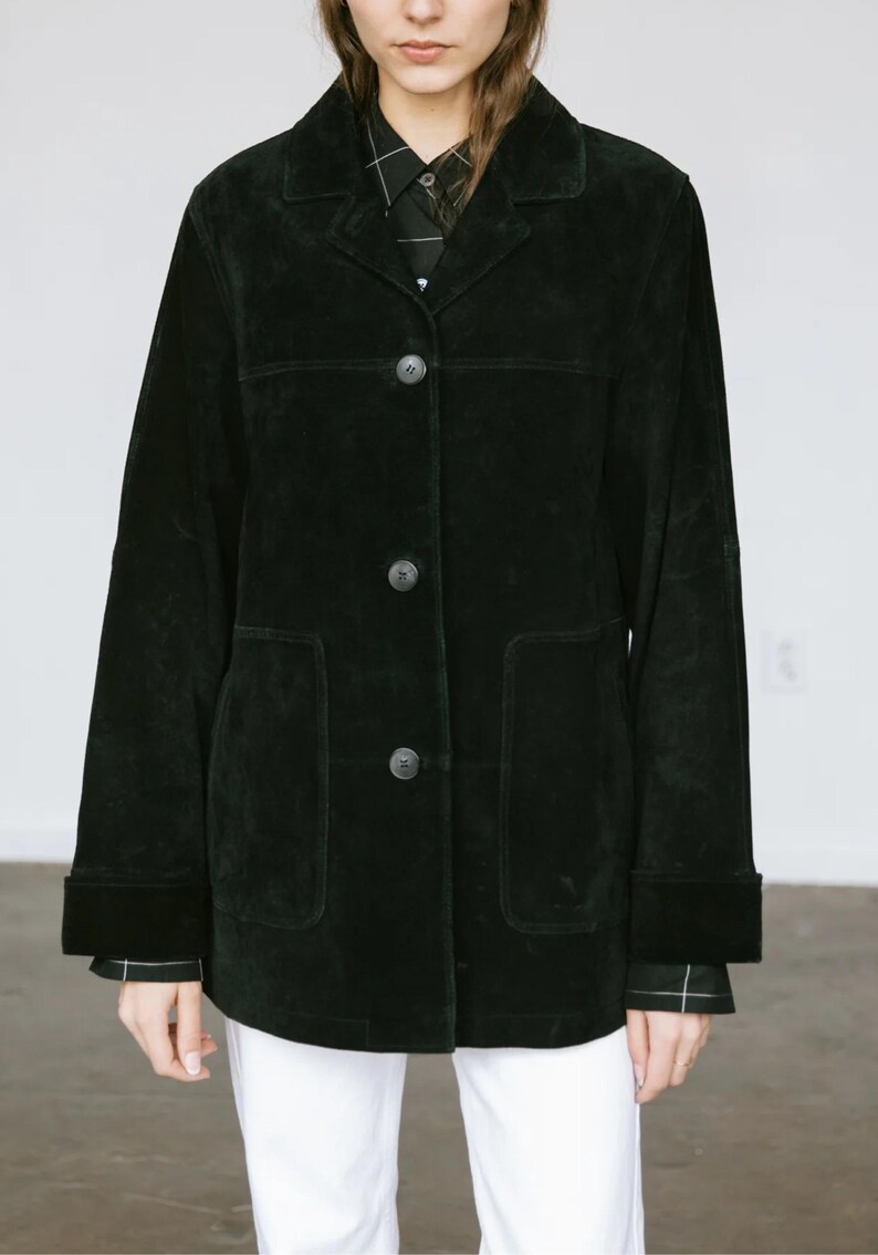 May include: A black suede jacket with a classic design. The jacket features a collared neckline, three buttons, and two front pockets. The sleeves have a cuff detail. The model is wearing a black and white patterned shirt underneath.