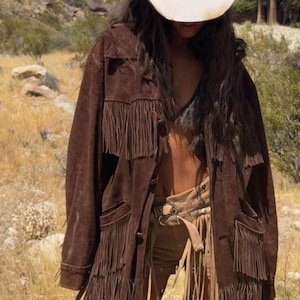 May include: Brown suede fringe jacket with a matching belt and shorts. The outfit is paired with a white cowboy hat. The jacket has pockets and button closures. The outfit is suitable for a western or bohemian theme.