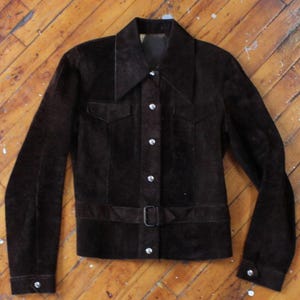 May include: A dark brown suede jacket with a collared neckline, two chest pockets, and a belt at the waist. The jacket has long sleeves with buttoned cuffs and a button-up front. The buttons are silver-colored.