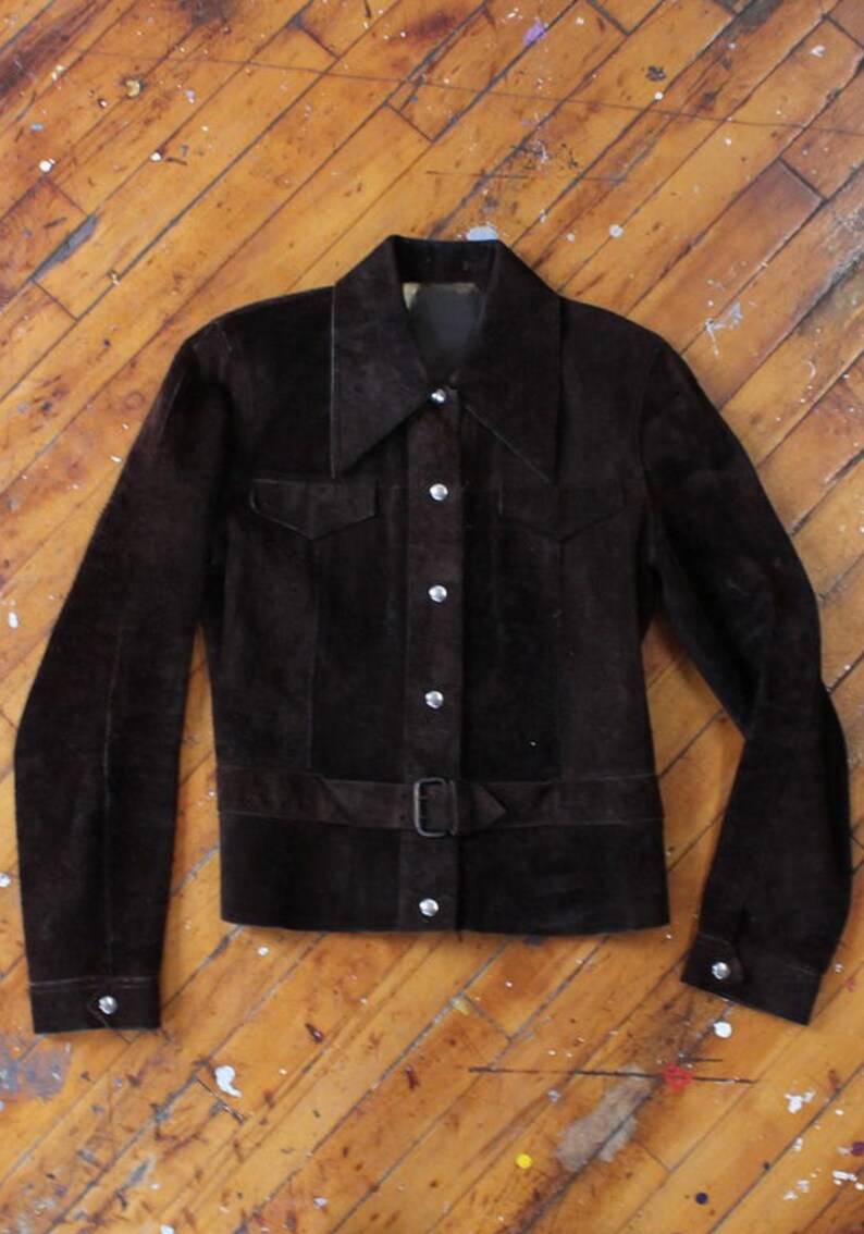 May include: A dark brown suede jacket with a collared neckline, two chest pockets, and a belt at the waist. The jacket has long sleeves with buttoned cuffs and a button-up front. The buttons are silver-colored.