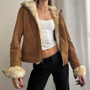 May include: A brown suede jacket with a faux fur-lined hood and cuffs. The jacket has a zipper closure and a light-colored fur lining. The sleeves and cuffs are trimmed with the same faux fur. The jacket is worn over a white tank top and black pinstripe pants.