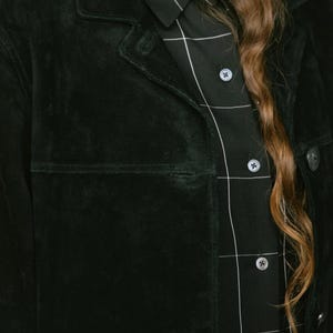 May include: A dark green suede jacket with a classic collar and button closure. The jacket is paired with a black button-down shirt featuring a white grid pattern. Long, wavy brown hair is visible.