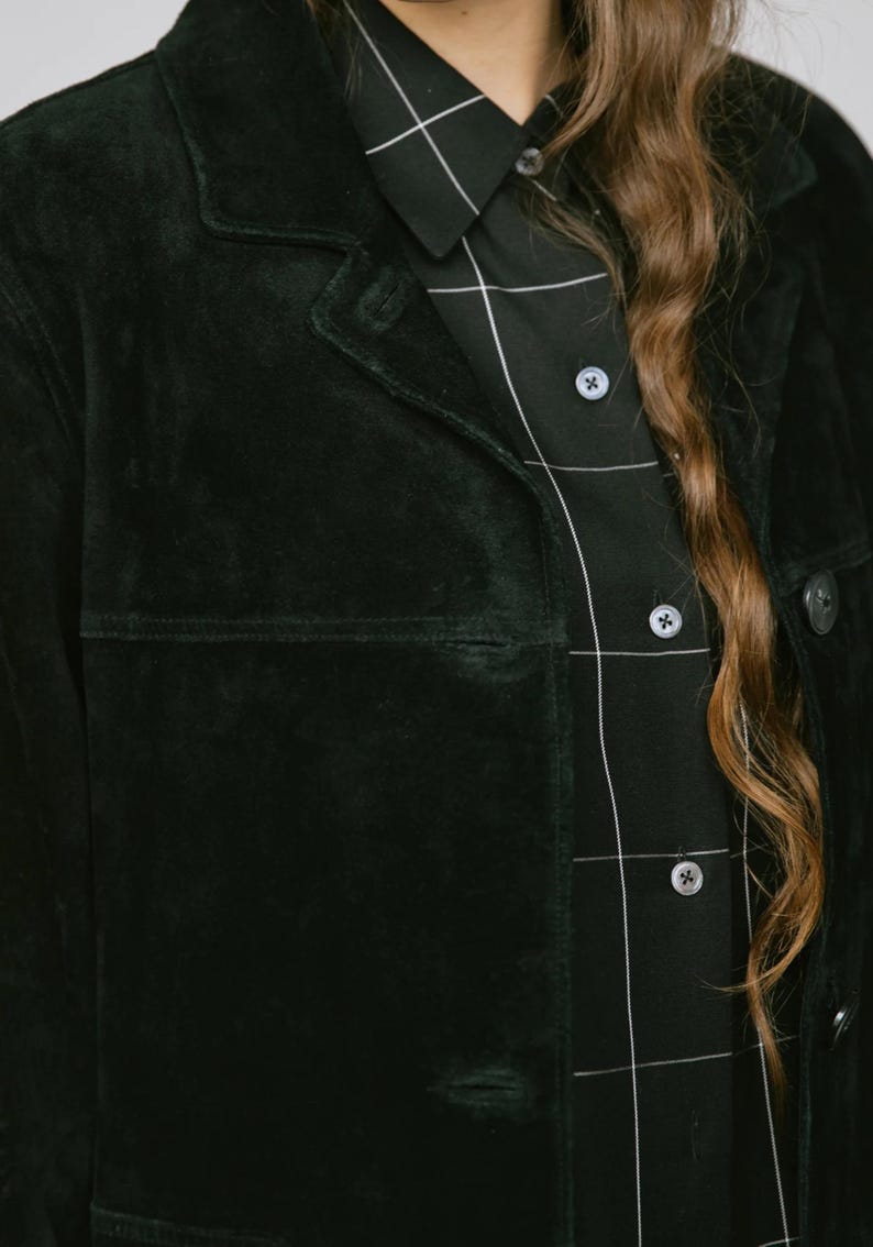 May include: A dark green suede jacket with a classic collar and button closure. The jacket is paired with a black button-down shirt featuring a white grid pattern. Long, wavy brown hair is visible.