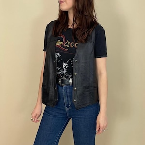 May include: Black leather vest with silver-tone buttons and pockets. Worn over a black graphic t-shirt and blue jeans. The t-shirt features a band logo and a silver-tone belt.