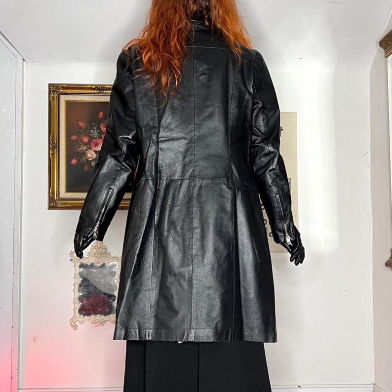 May include: A black leather coat with a tailored fit, featuring a notched collar, zipper details on the sleeves, and a pleated skirt. The coat is knee-length and appears to be vintage or vintage-inspired.