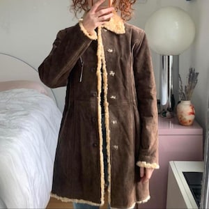 May include: A brown suede coat with a faux fur collar and trim. The coat has a button-up front and a long, tailored silhouette. The interior lining is a light tan color.