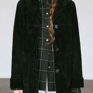May include: A black suede jacket with a button-up front and two front pockets. The jacket is worn over a black and white plaid shirt. The sleeves are long, and the jacket has a classic, tailored look. The model is wearing white pants.