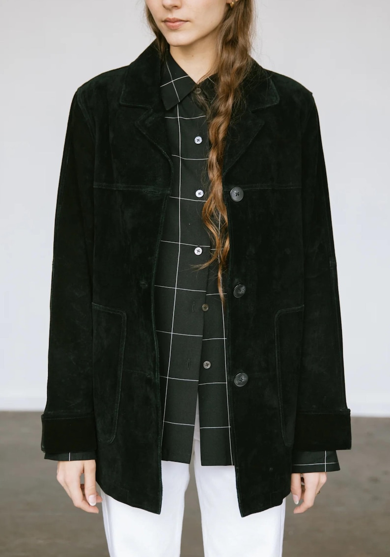 May include: A black suede jacket with a button-up front and two front pockets. The jacket is worn over a black and white plaid shirt. The sleeves are long, and the jacket has a classic, tailored look. The model is wearing white pants.