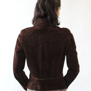 May include: A dark brown suede jacket with a high collar and a belt detail at the waist. The jacket has long sleeves and a tailored fit, showcasing a classic and elegant design. The back view highlights the jacket's structured silhouette.