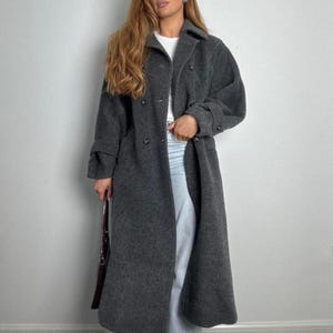 May include: A long, charcoal gray coat with a double-breasted design. The coat features a classic collar, button closures, and long sleeves with buttoned cuffs. The coat is knee-length and appears to be made of a wool blend.