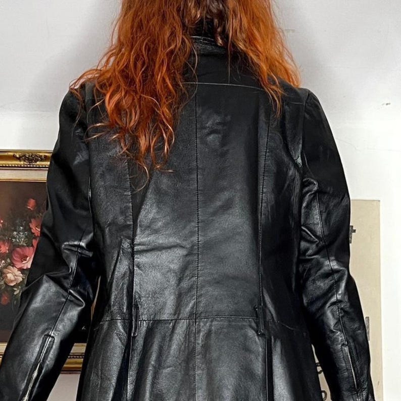 May include: A black leather jacket with a tailored fit. The jacket has a high collar, long sleeves, and a button-down front. The back of the jacket features vertical seams and a slight flare at the waist. The jacket is worn by a person with red hair.