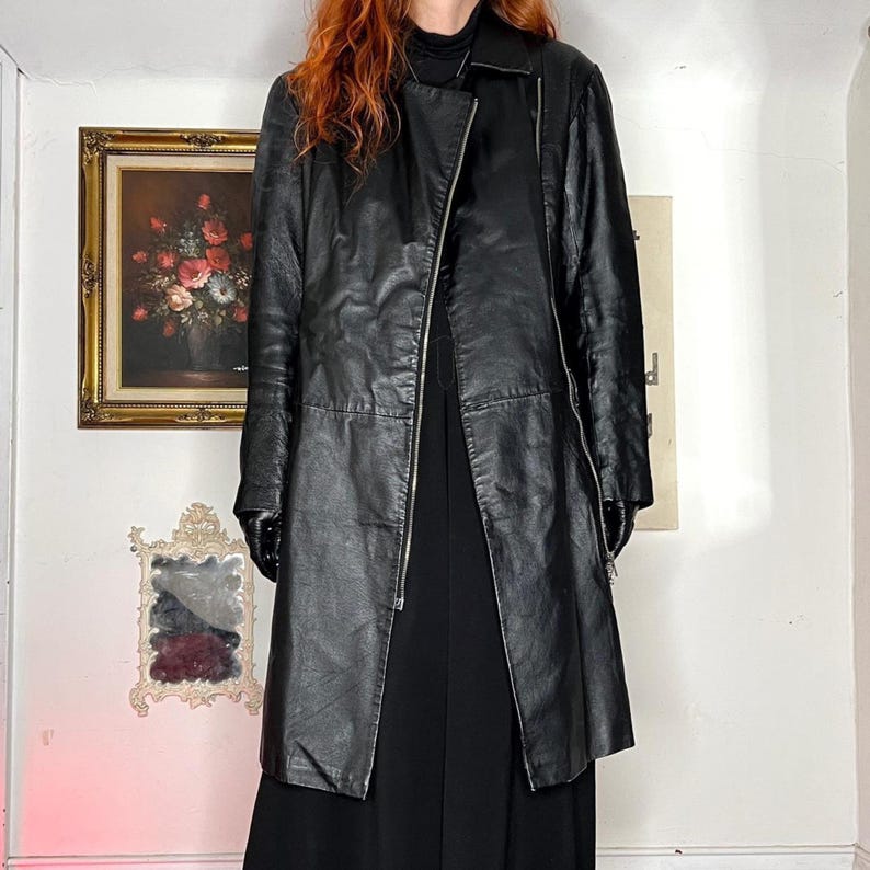 May include: A black leather coat with a zip fastening. The coat is knee-length and has a classic design. The coat is worn over a black outfit. The background includes a painting and a mirror.