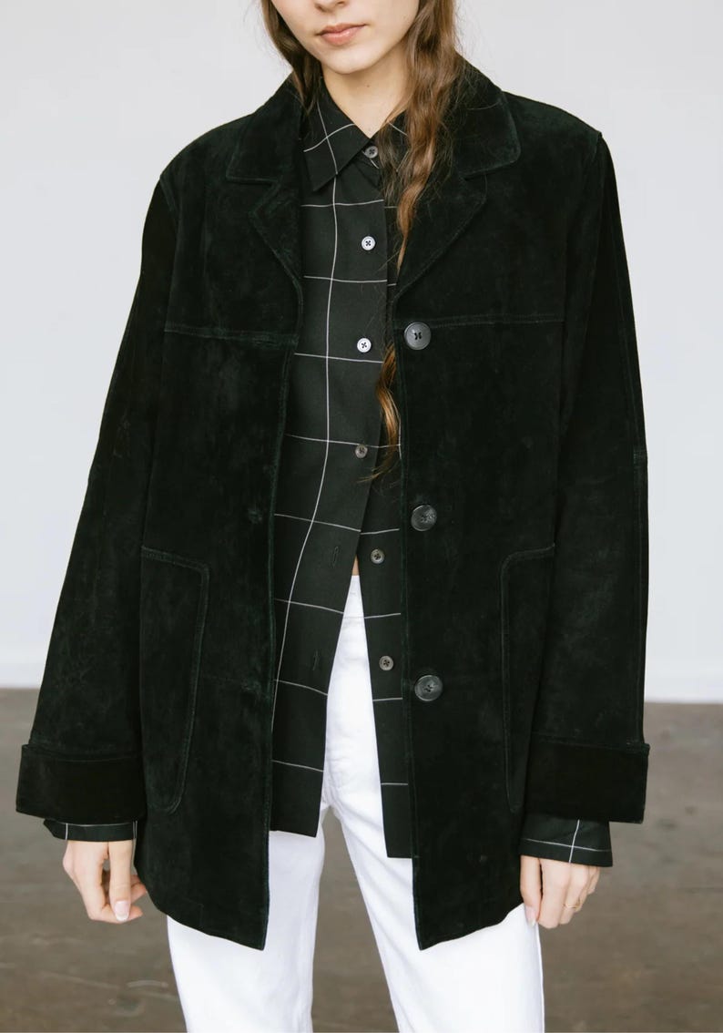 May include: A black suede jacket with a classic collar and button closure. The jacket has two front pockets and long sleeves with folded cuffs. Underneath, a black and white plaid shirt is visible, paired with white pants.
