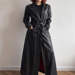 May include: A long black leather trench coat featuring a notched lapel collar, long sleeves, and a matching tie belt at the waist. The coat has two front pockets and a floor-length hemline, worn over a plain black shirt and black square-toe boots.