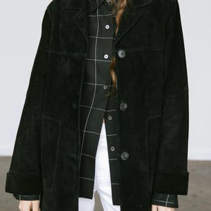 May include: A black suede jacket with a button-up front, worn over a black and white plaid shirt and white pants. The jacket has a classic collar and two front pockets. The sleeves are cuffed at the wrists.