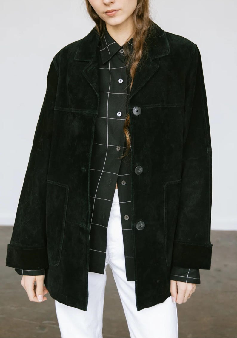 May include: A black suede jacket with a button-up front, worn over a black and white plaid shirt and white pants. The jacket has a classic collar and two front pockets. The sleeves are cuffed at the wrists.