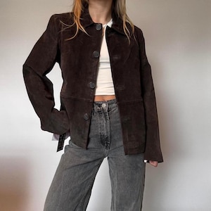 May include: A dark brown suede jacket with a button-up front and a collared neckline. The jacket has long sleeves and a tailored fit. The model is wearing the jacket with a white top and gray jeans.
