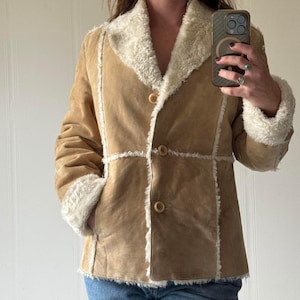 May include: A tan suede jacket with a cream-colored faux fur collar and trim. The jacket has a button-up front and long sleeves with fur cuffs. The jacket is a classic style, perfect for layering in cooler weather.