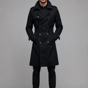 May include: A black double-breasted trench coat featuring a wide collar, shoulder epaulets, and a matching waist belt with a buckle. The coat has long sleeves with buckled cuffs, button closures, and a knee-length hem, worn over black trousers and boots.