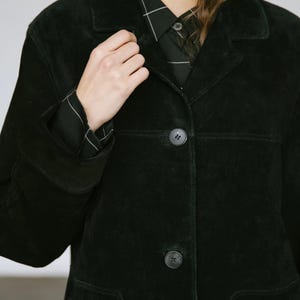 May include: A black suede jacket with a classic collar and button closure. The jacket features a tailored fit, with visible seams and pockets. The sleeves are long, and the buttons are dark. The jacket is worn over a black and white checkered shirt.