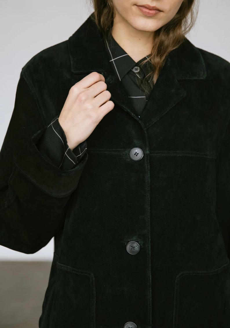 May include: A black suede jacket with a classic collar and button closure. The jacket features a tailored fit, with visible seams and pockets. The sleeves are long, and the buttons are dark. The jacket is worn over a black and white checkered shirt.