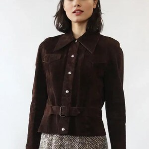 May include: A dark brown suede jacket with a collared neckline, button-up front, and a belt at the waist. The jacket has long sleeves and two small pockets. The model is wearing a patterned skirt.