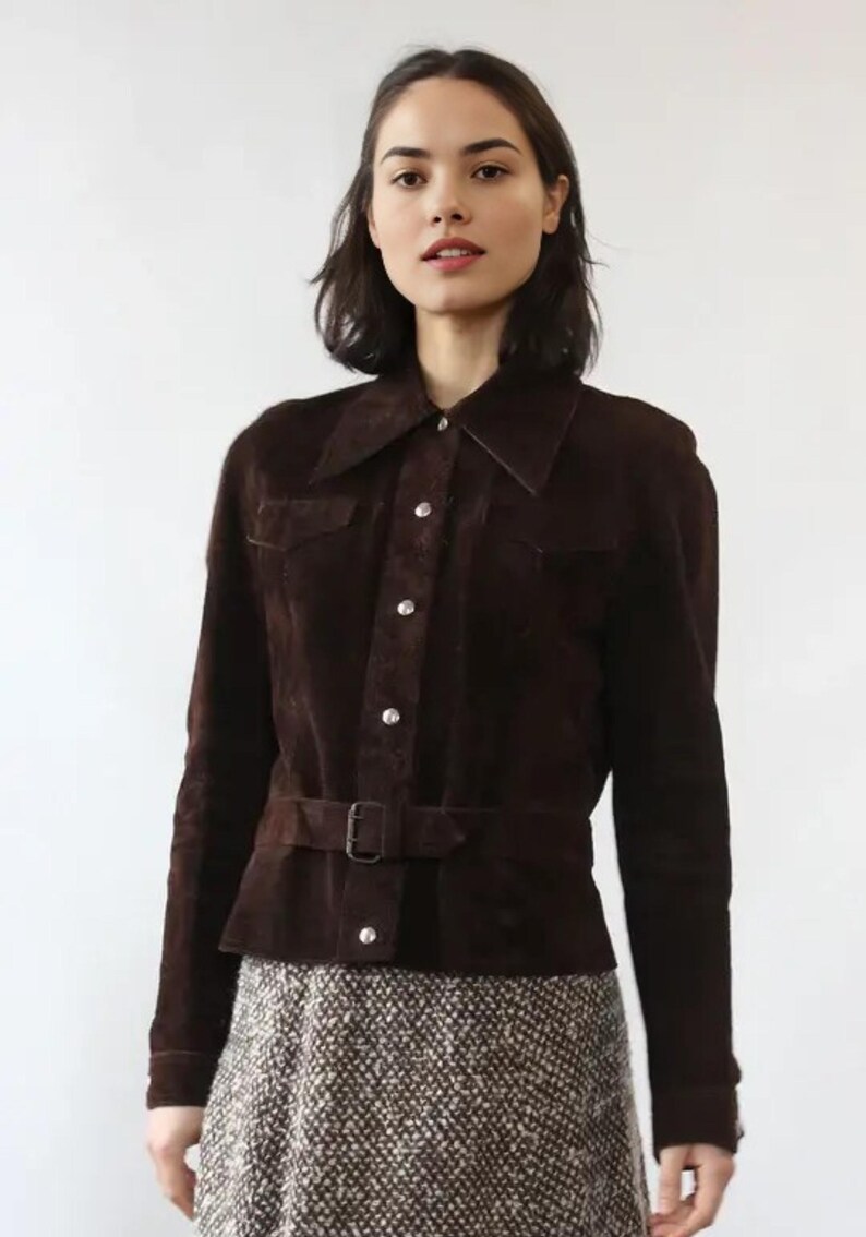 May include: A dark brown suede jacket with a collared neckline, button-up front, and a belt at the waist. The jacket has long sleeves and two small pockets. The model is wearing a patterned skirt.
