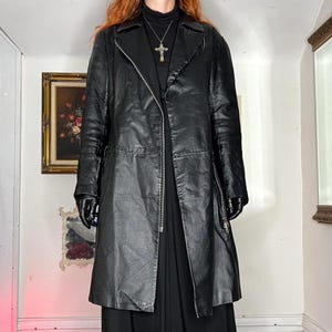 May include: A long, black leather coat with a zip fastening. The coat features a classic lapel collar and a tailored fit. The sleeves are long, and the coat extends to mid-calf length. The coat is worn with black gloves and a black dress.