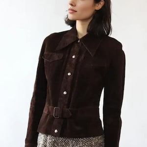 May include: A dark brown suede jacket with a collared neckline, button-up front, and a matching belt. The jacket features two chest pockets and long sleeves. The model is wearing a patterned skirt.