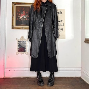 May include: A black leather coat with a zip fastening, worn over a black dress. The coat features a classic design with a lapel collar. The outfit is completed with black boots. The background includes framed artwork and a sign.