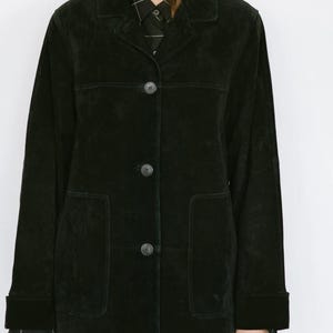 May include: Black suede jacket with a classic collar, three-button closure, and two front pockets. The jacket has long sleeves with buttoned cuffs. The model is wearing a black and white patterned shirt underneath.