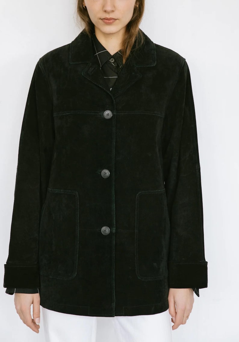 May include: Black suede jacket with a classic collar, three-button closure, and two front pockets. The jacket has long sleeves with buttoned cuffs. The model is wearing a black and white patterned shirt underneath.