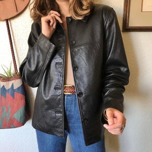 Peut inclure: Veste en cuir noire à boutons. Le modèle porte un jean bleu et une ceinture marron. La veste a un design classique avec un col et des manches longues. La veste semble être en bon état.